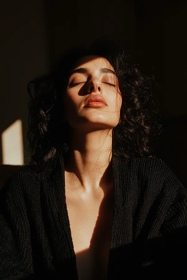Close-up portrait of a woman with curly hair and eyes closed, lit by warm sunlight against a dark background, wearing a black textured robe.