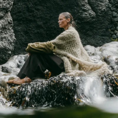 Woman in layered neutral knitwear seated beside a stream among dark coastal rocks.