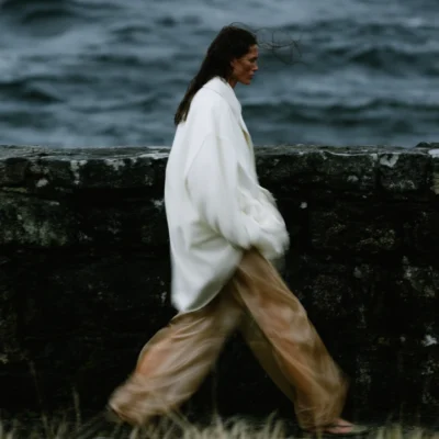 Woman in a white oversized jacket and flowing neutral trousers walking beside a dark sea wall.