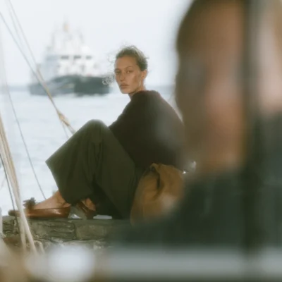 Woman in black clothing seated beside harbour water with a boat softly visible behind her.