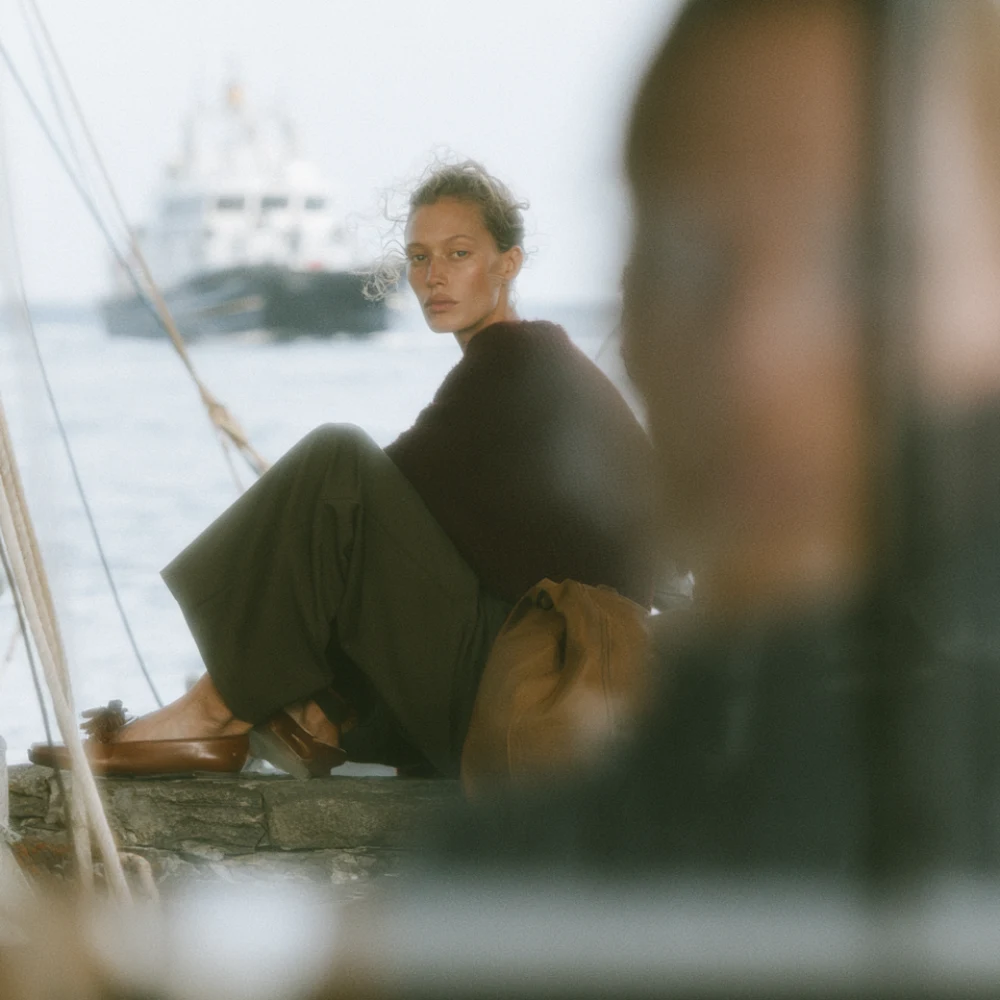 Woman in black clothing seated beside harbour water with a boat softly visible behind her.