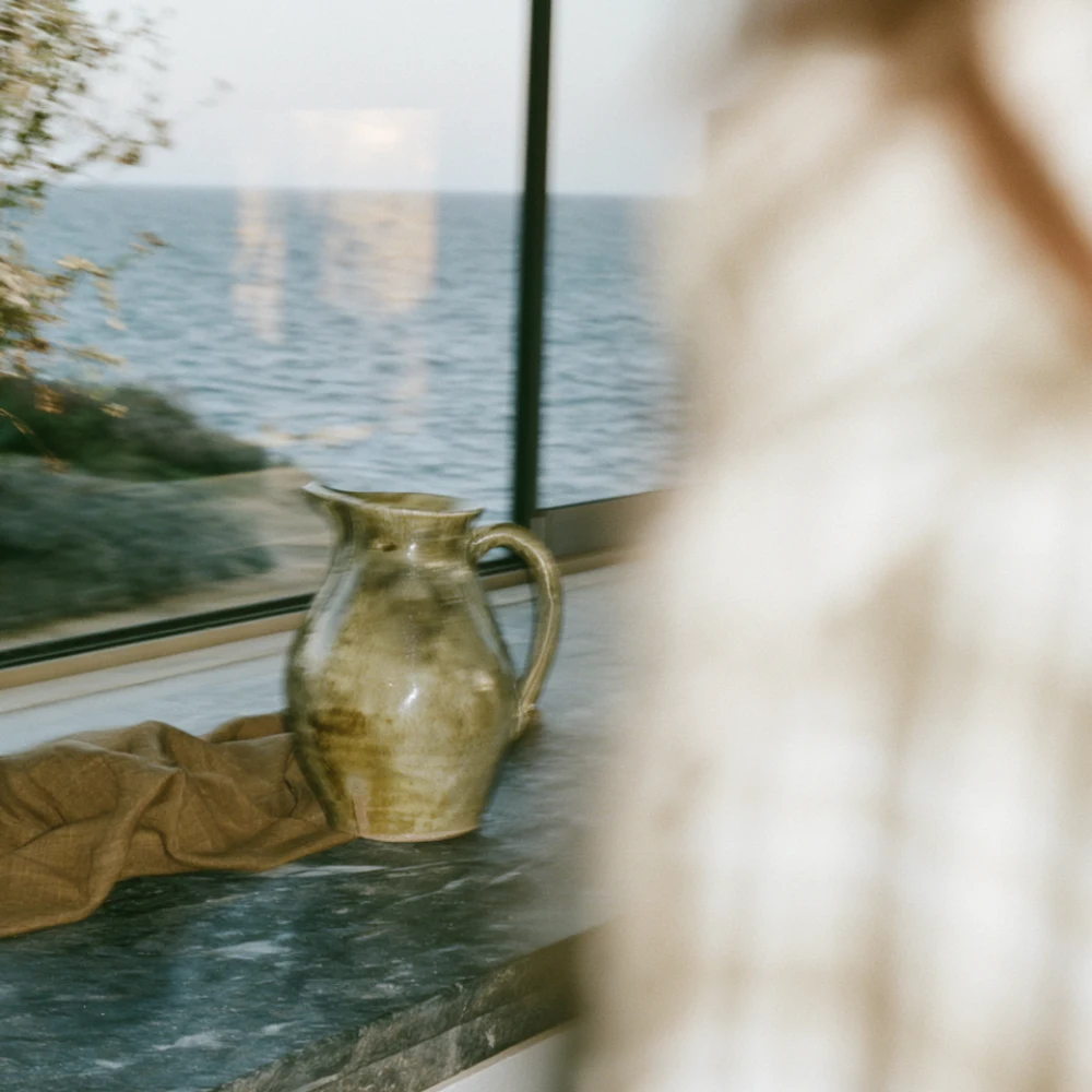 Green ceramic pitcher and folded brown fabric on a stone counter overlooking the sea.