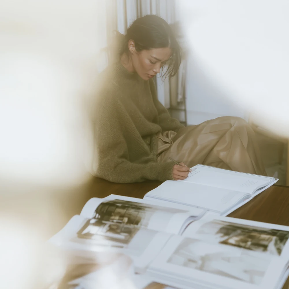 Woman writing in a notebook beside open art books in soft window light.